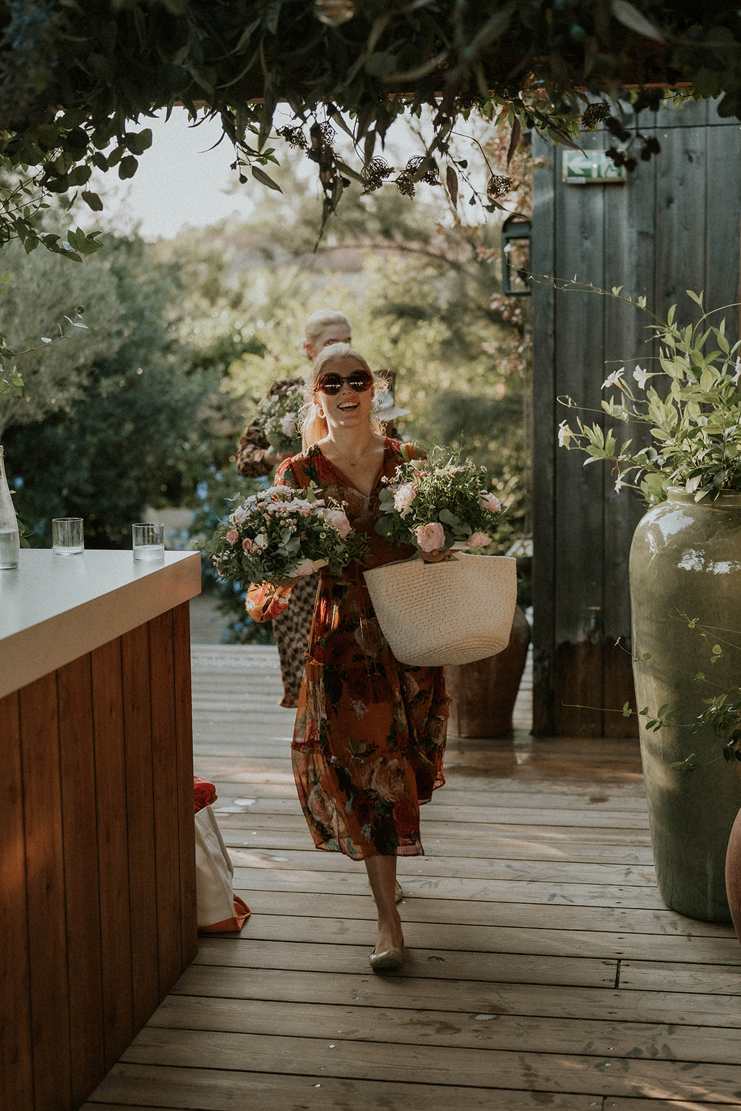 Kalu laughing in sunglasses and a floral dress, carrying bouquets of pink roses across a wooden terrace under olive branches.