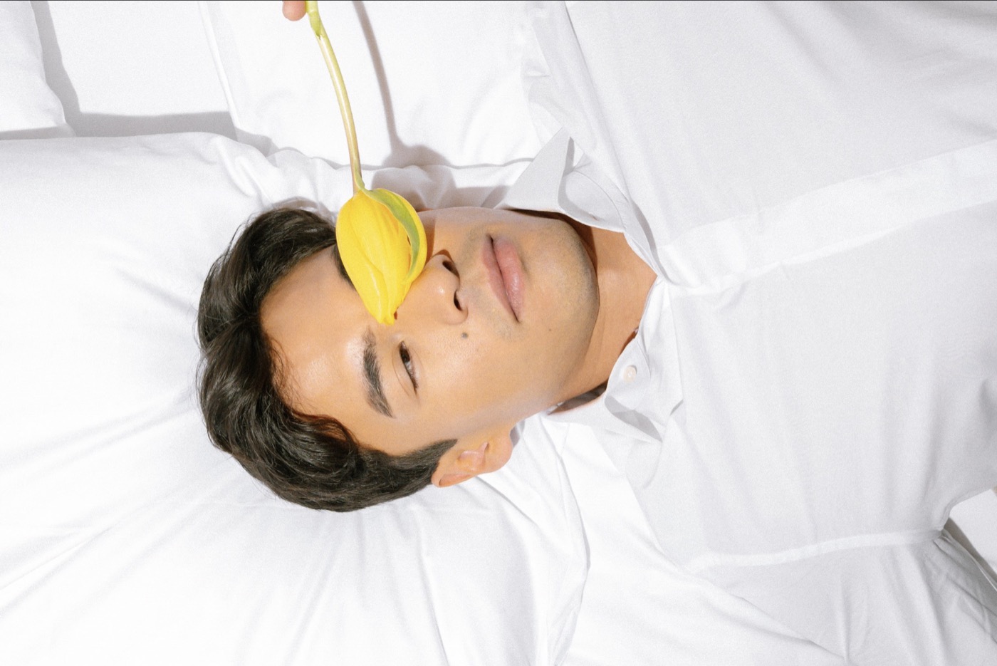 Man in a white shirt lying on white bedding, a yellow tulip held over one eye