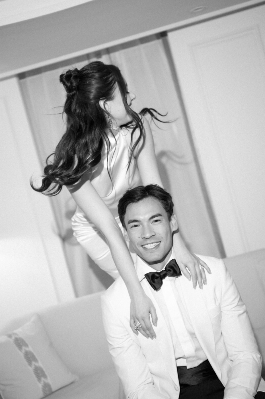 Black-and-white photo of a smiling groom in a white dinner jacket, the bride leaning over him with her hair flying
