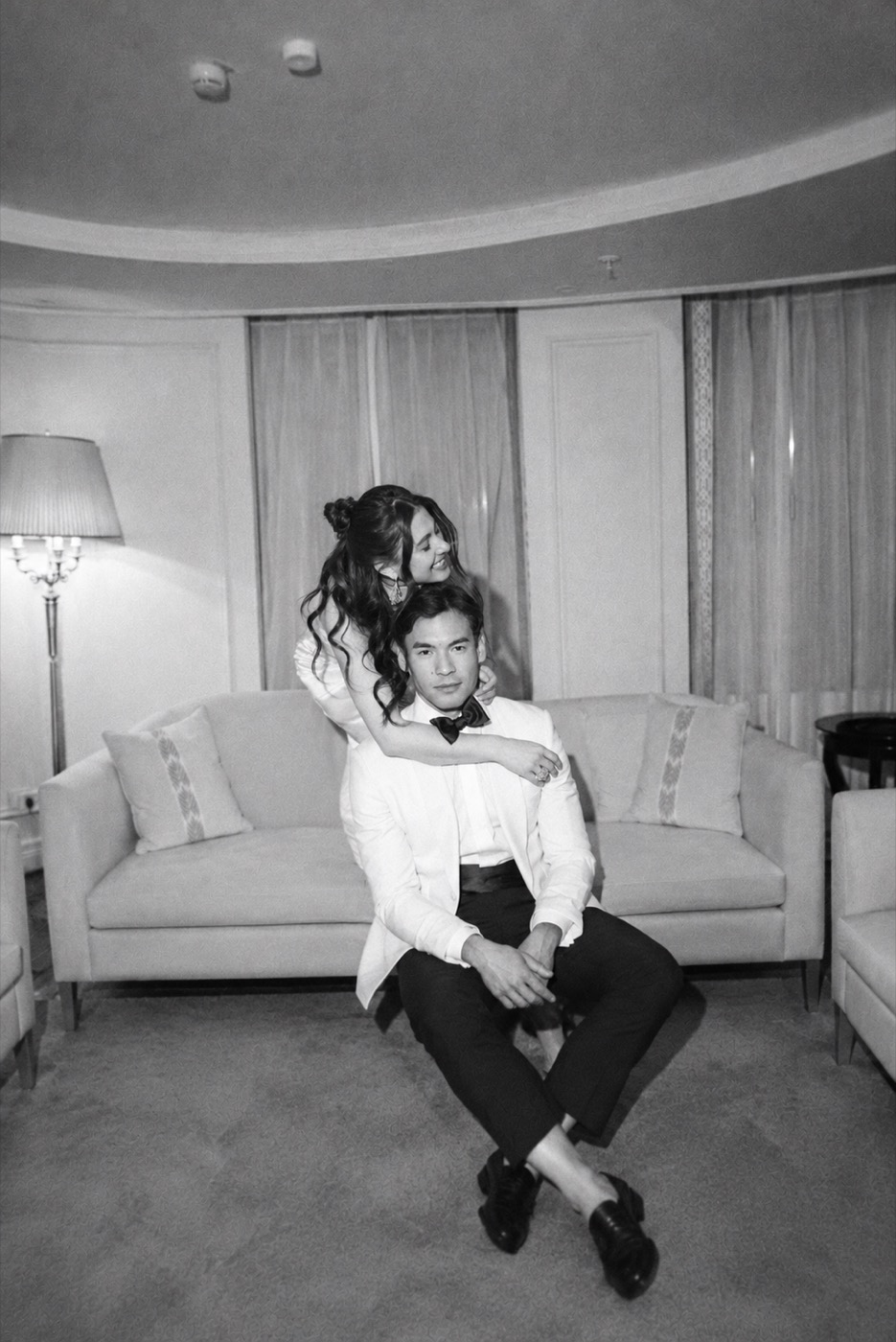Black-and-white photo of a bride hugging her groom from behind in front of an elegant sofa